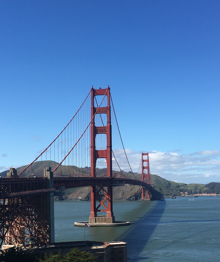 Photo of Golden Gate Bridge on sunny day with water in foreground, buildings in lower right, hills and clouds in background.