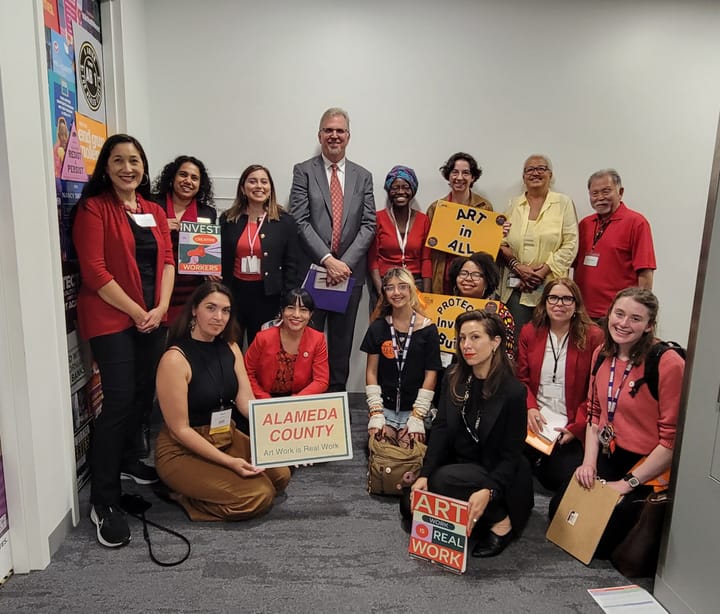 14 arts advocates, some with signs supporting arts workers, flank State Senator Nancy Skinner’s policy advisor Robert Gammon